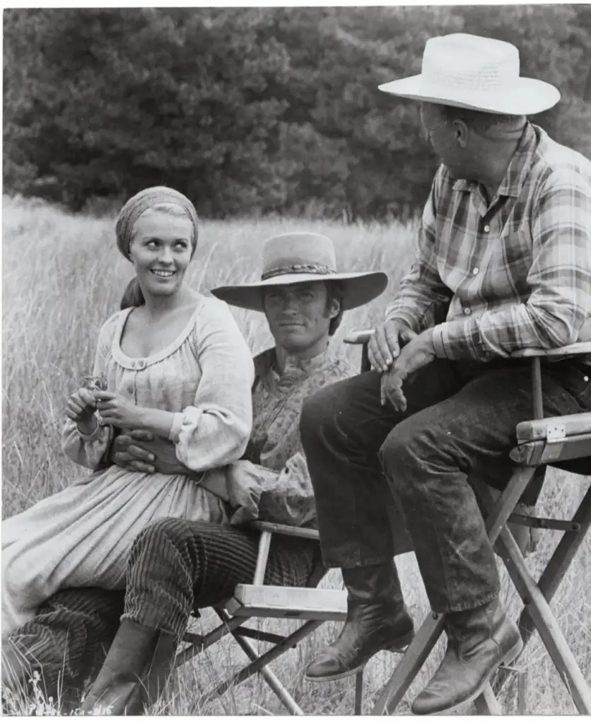 Three people sit outdoors. A woman in a headscarf smiles while sitting next to a man in a cowboy hat. Another man in a plaid shirt and hat sits to the right, looking at them. They are in a grassy field with trees in the background.