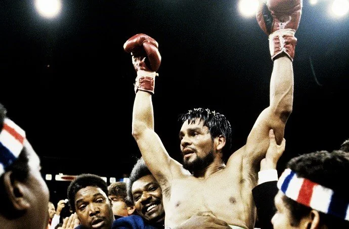 A triumphant boxer with red gloves is lifted by a crowd in a boxing ring. He has one arm raised high, and the scene is filled with cheering spectators. The background is brightly lit, emphasizing the victorious moment.