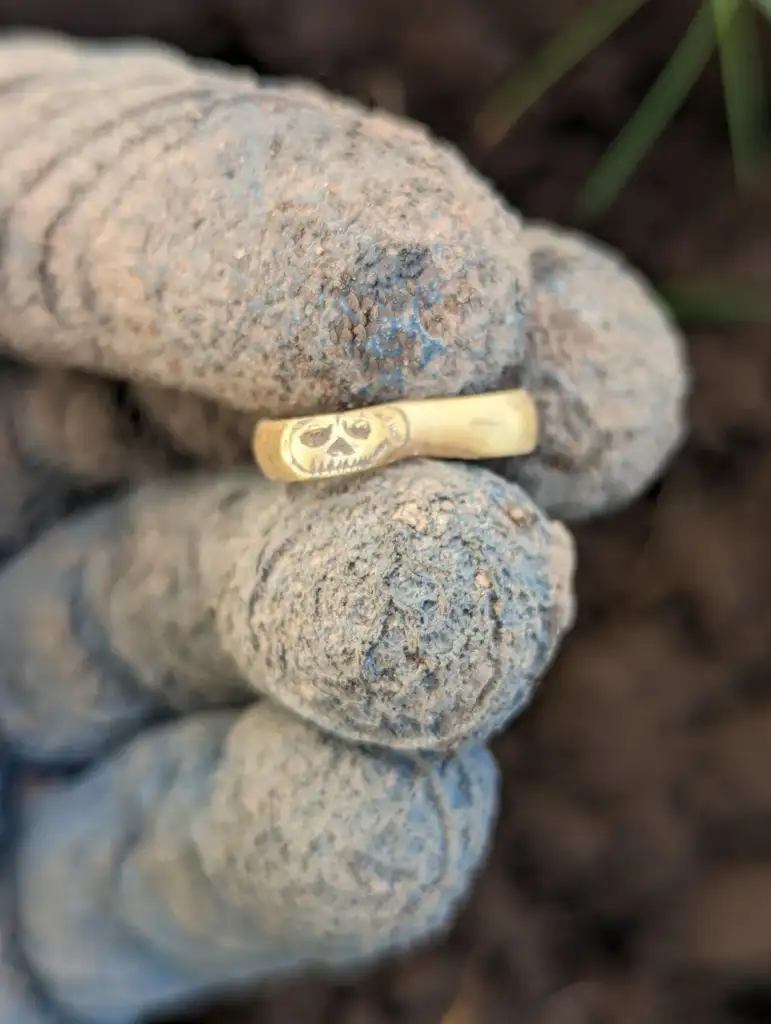 A muddy hand holds a gold ring engraved with a skull design. The background is blurred soil.