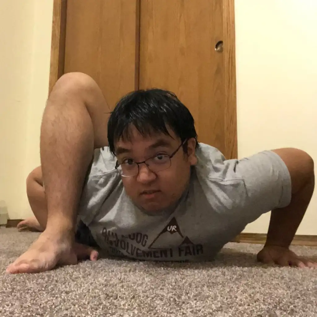 A person in a gray t-shirt is performing an advanced yoga pose on a carpeted floor. They are balancing on their arms with legs wrapped over their shoulders, in front of a wooden door.