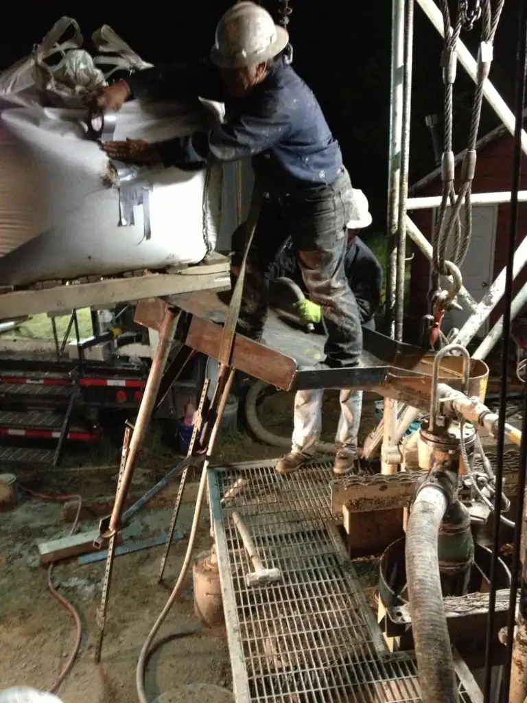 Two construction workers at a work site. One stands on a metal grate, securing a large bag suspended by a crane, while the other supports nearby. Equipment and structural components surround the scene at night.