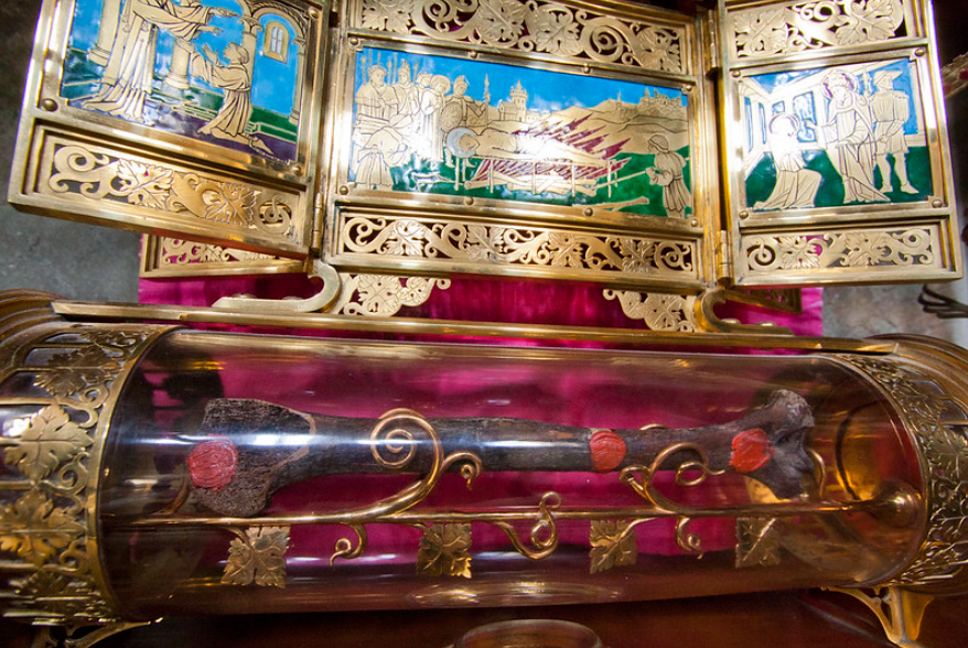 A glass and gold reliquary containing a relic is displayed. The background features intricate gold designs and colorful stained glass art depicting historical scenes.