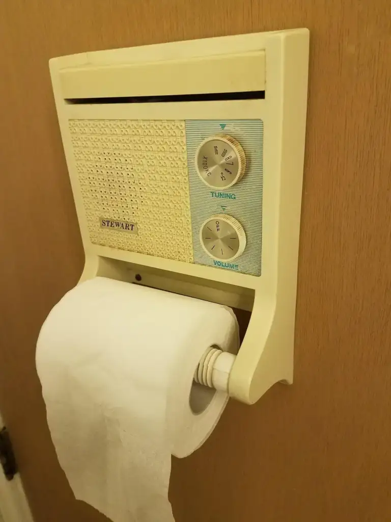 A vintage wall-mounted radio with a tuning and volume dial is installed above a toilet paper holder. The radio's brand "Stewart" is visible on the front panel. A roll of toilet paper hangs from the holder below.