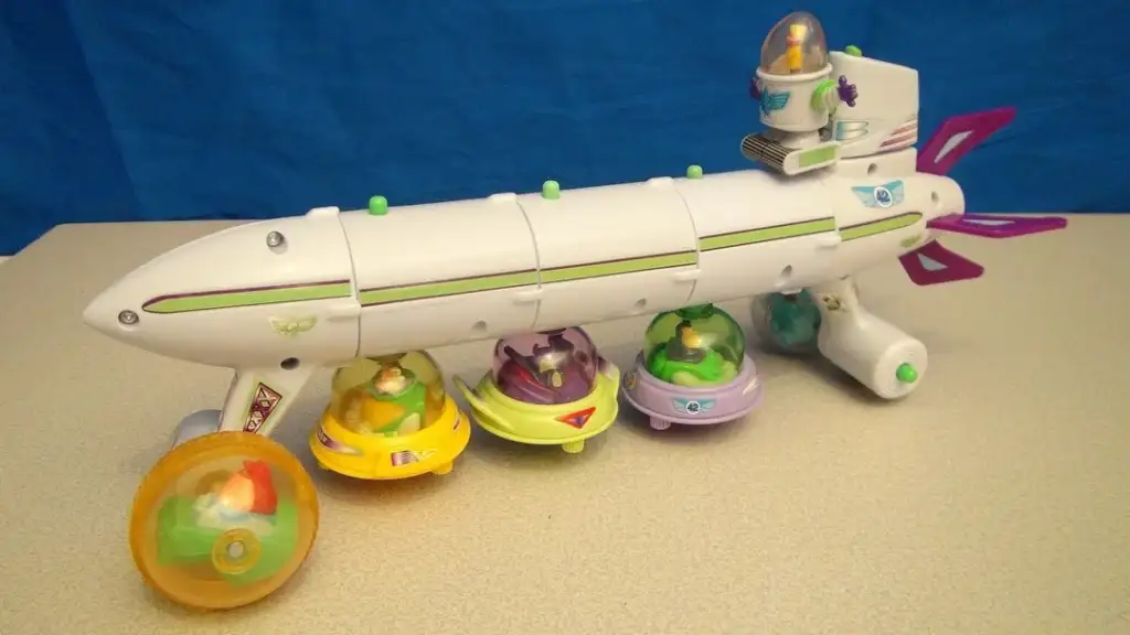 Toy spaceship with a white body, purple accents, and a propeller at the rear. It sits on colorful wheels, each with animal figures inside plastic domes. A small character is visible on top of the spaceship, set against a blue background.