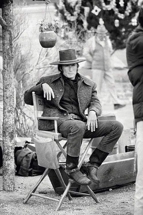 Black and white image of a person in a cowboy outfit sitting on a director's chair outdoors. They wear a wide-brimmed hat, dark jacket, and boots, with a relaxed pose. There’s a hanging pot above and blurred people in the background.