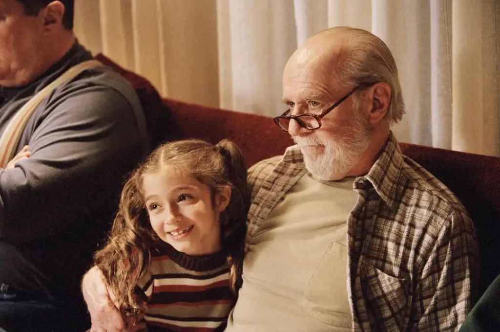 An elderly man with a white beard and glasses sits on a couch, smiling gently. He has his arm around a young girl with wavy hair in pigtails, who is also smiling. They both appear content and relaxed in a cozy setting.