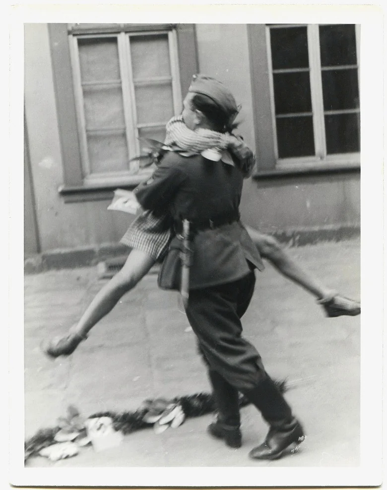 A soldier joyfully carries a woman in a striped dress down a cobblestone street. Both appear excited, with the woman lifted off the ground. The buildings in the background have a few windows. A small plant lies on the street.