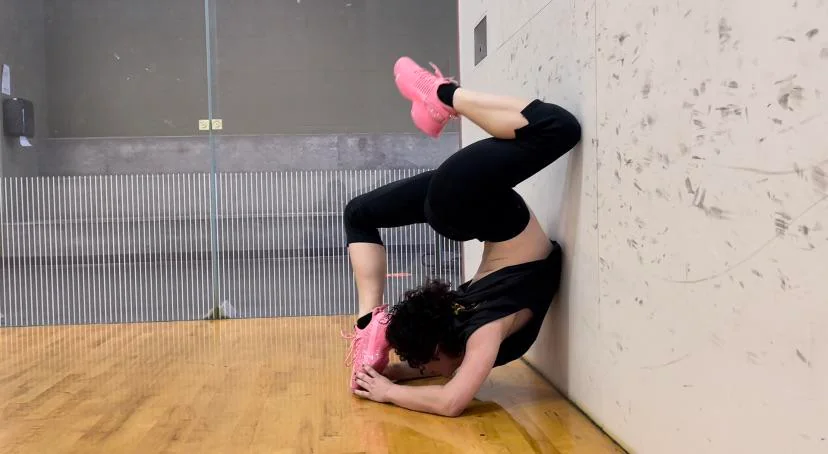 A person performs a headstand against a wall in a gym, wearing black athletic wear and bright pink shoes. The room has a wooden floor and visible glass walls on the side.