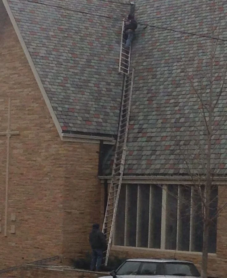 Two individuals are using an extended ladder to work on a steep church roof. One person is on the roof, while the other stands at the base. A tree and a parked car are visible nearby.