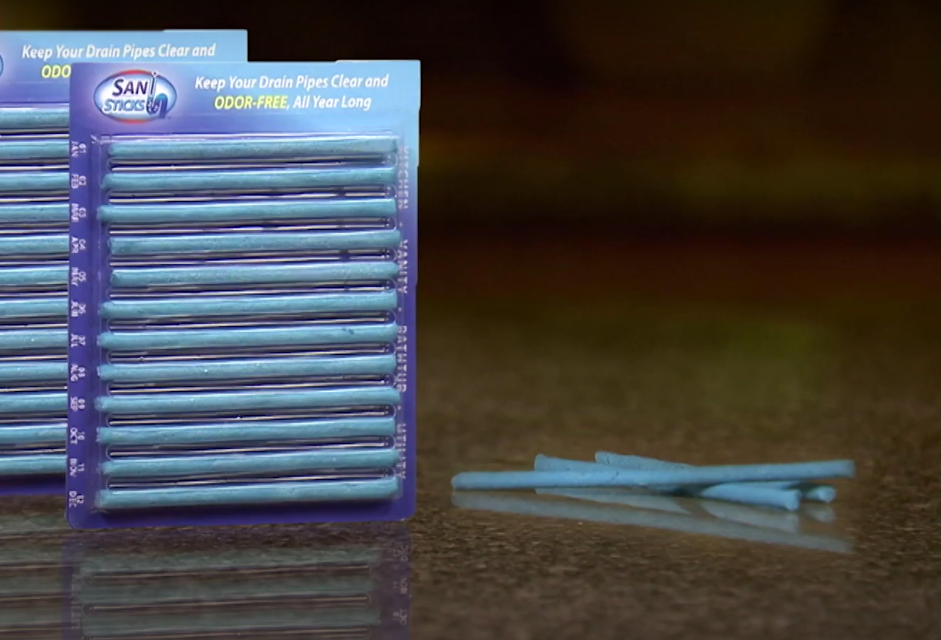 The image shows two packages of blue drain sticks labeled "SAN STICKS" with some sticks laid out in front. The packaging claims to keep drain pipes clear and odor-free. They are displayed on a reflective surface.