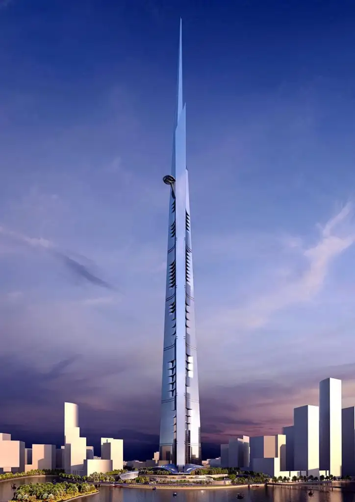 A futuristic skyscraper with a sleek, tapering design stands tall against a dusk sky. Surrounded by low-rise buildings and greenery, the structure features vertical lines and a pointed top, conveying a sense of modern architectural innovation.