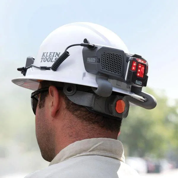 A person wearing a white hard hat with "Klein Tools" branding. Attached to the helmet is a device with red lights and a cord. The person is outdoors, wearing sunglasses and looking away.