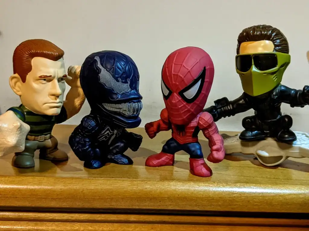 Four stylized action figures are displayed on a wooden surface. From left to right: a muscular figure with a serious expression, a dark figure with a toothy grin, a red and blue figure with web patterns, and a figure in sunglasses and a green scarf.