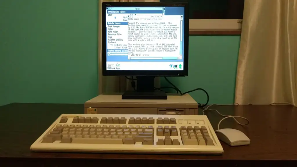 A vintage NEC desktop computer is set up on a wooden desk. The setup includes a CRT monitor displaying a text-based program, a beige keyboard, and a gray mouse. The background features a window with a curtain and a teal wall.