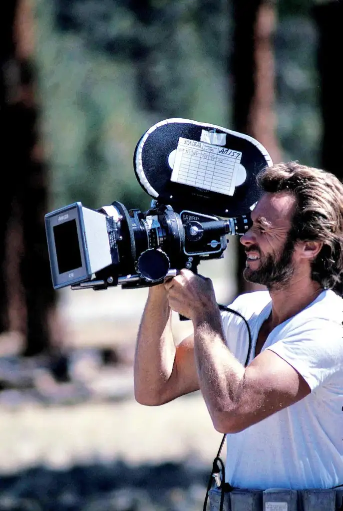 A man with a beard and mustache is outdoors, operating a vintage film camera mounted on his shoulder. He wears a white T-shirt and looks through the camera's viewfinder, appearing focused. The background is blurred with trees.