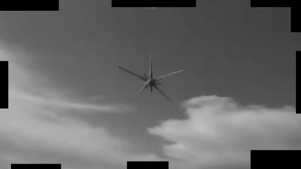 A black and white image of a helicopter viewed from below, set against a partly cloudy sky. The camera is focused on the rotor blades spinning above the helicopter. Black borders frame the edges of the image.