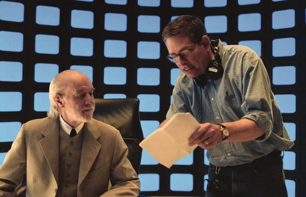 An older man with a beard in a beige suit sits at a desk, looking at a younger man in glasses standing next to him, who is holding and pointing at a paper. The background has a grid pattern with a blue glow.