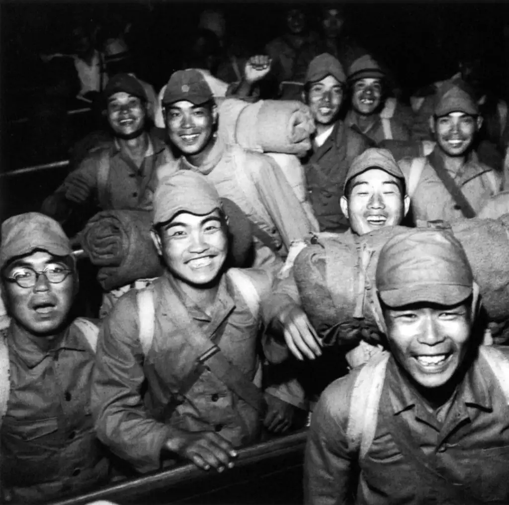 A group of smiling soldiers in uniform, carrying rolled blankets on their backs. The photo is black and white, and the soldiers appear joyful and excited, looking directly at the camera.