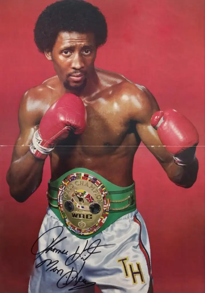 A boxer wearing red gloves and white shorts stands in a fighting stance. He has an "International WBC Champion" belt with a green strap. The initials "TH" are on his shorts. The background is red.