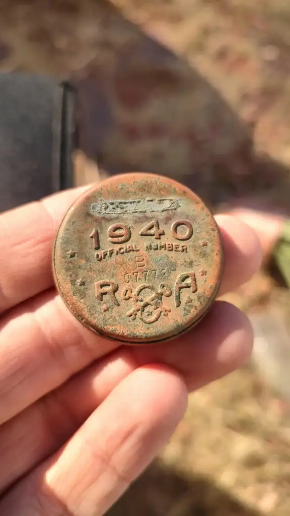 Close-up of a person's hand holding a round, worn badge with "1940 Official Member RCRA" and other illegible text. The badge has a rusty appearance and is held outdoors.