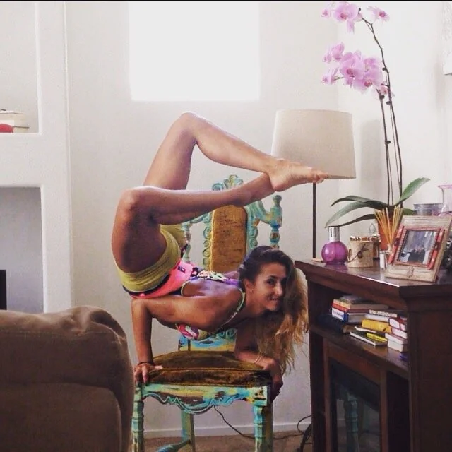 A person performs a yoga pose on a chair, balancing on forearms with legs arched over the head. The room has a lamp, books, and a potted orchid on a table. The person wears colorful workout clothes.