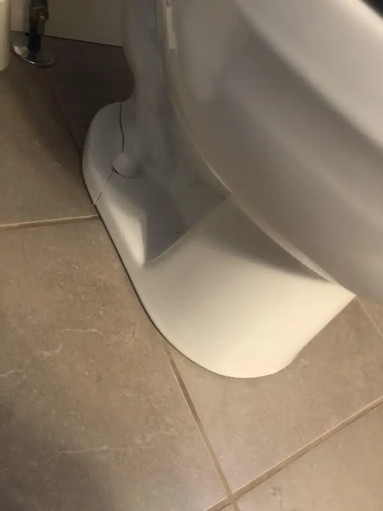 A close-up image of the base of a white toilet on a tiled floor. There is a visible crack running through the ceramic material near the floor joint.