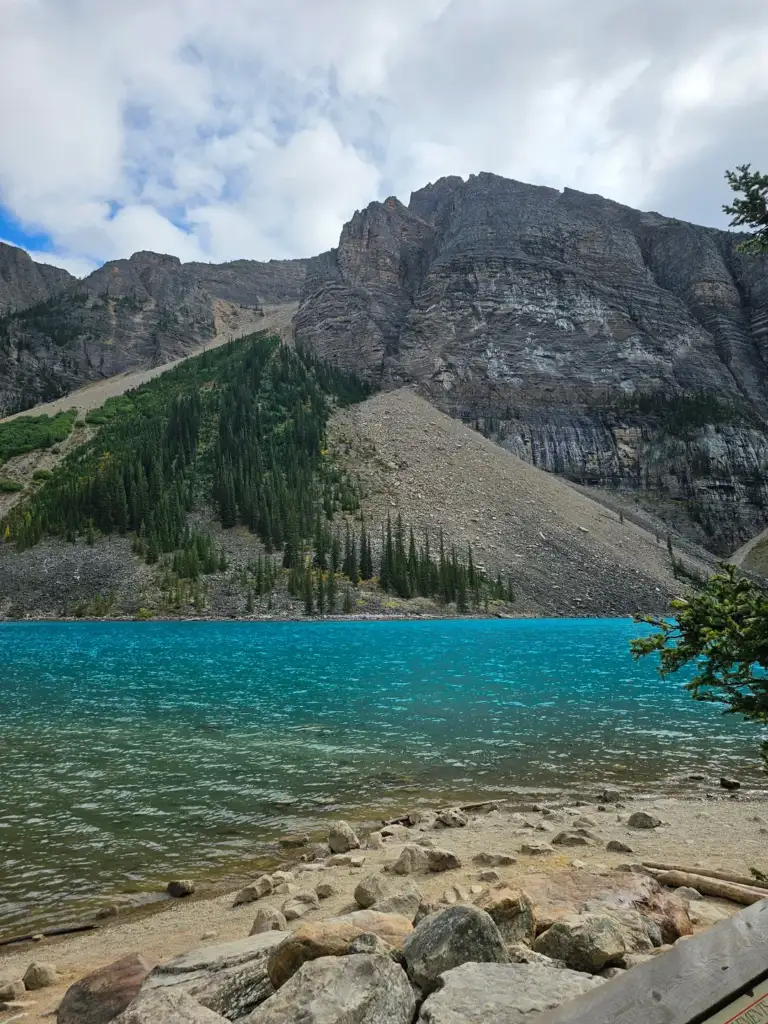 A vibrant turquoise lake bordered by rocky shores sits below a steep, towering mountain. Evergreen trees dot the lower slopes under a partly cloudy sky.