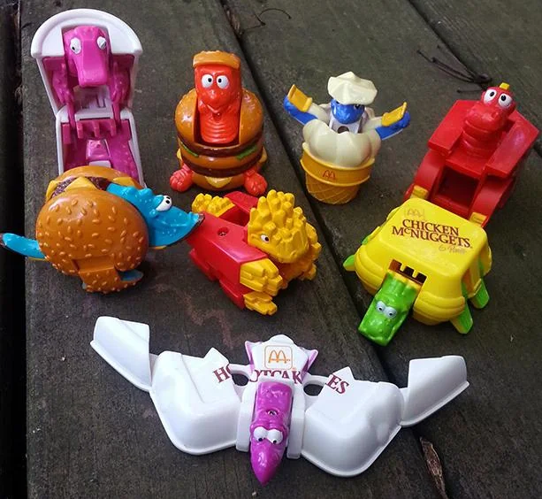 Assorted vintage McDonald's Happy Meal toys on a wooden surface. The toys are colorful, with designs resembling food items like chicken nuggets and pancakes featured in theme park ride shapes.