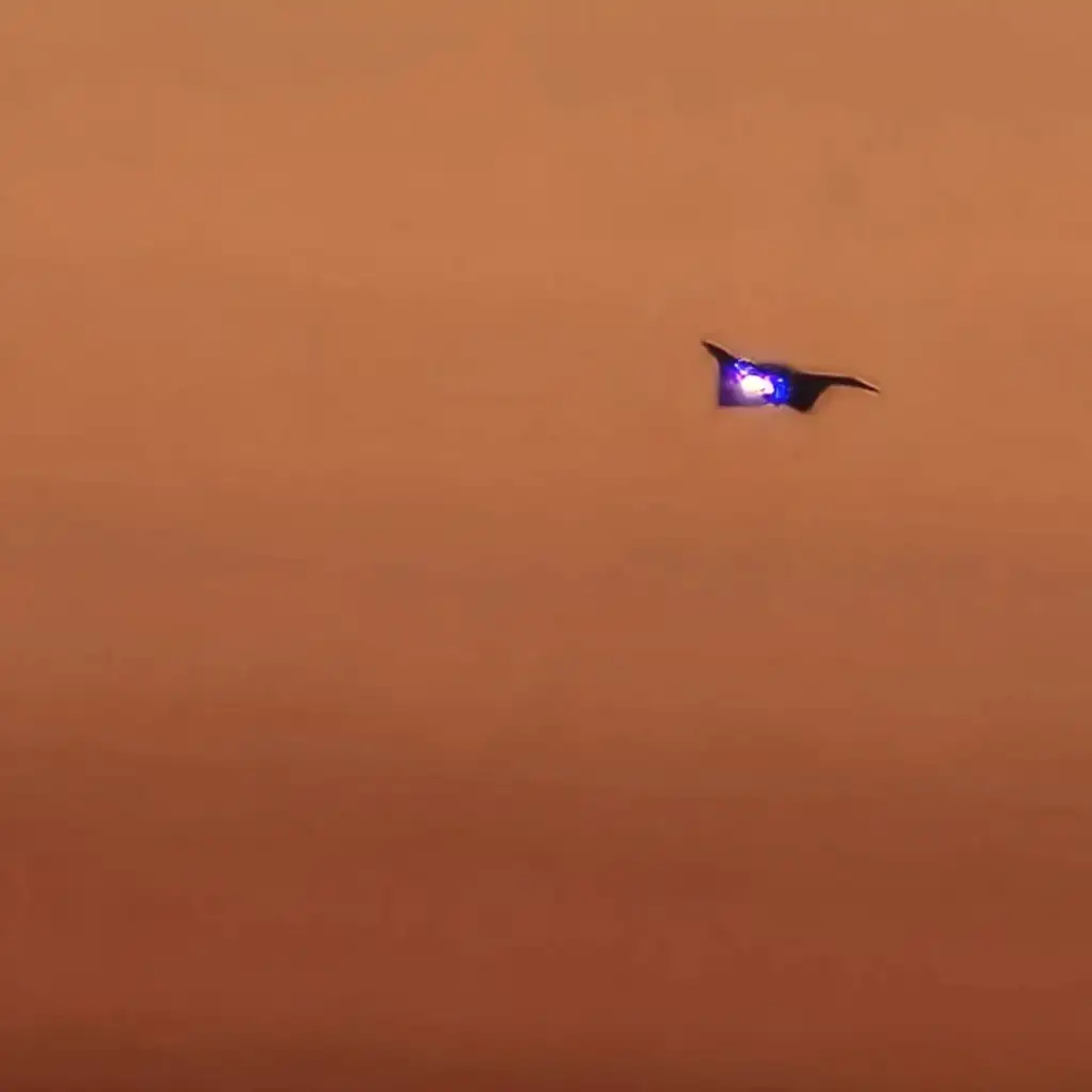 A drone with blue lights flying against a background of a hazy, orange-brown sky. The scene suggests dusk or a sunset, creating a mysterious and serene atmosphere.