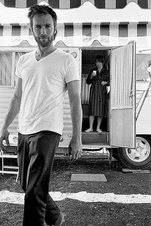 A man in a white T-shirt and dark pants walks towards the camera with a serious expression. In the background, a person in a robe stands at the doorway of a trailer, holding a mug. The trailer has a striped awning. The scene is in black and white.