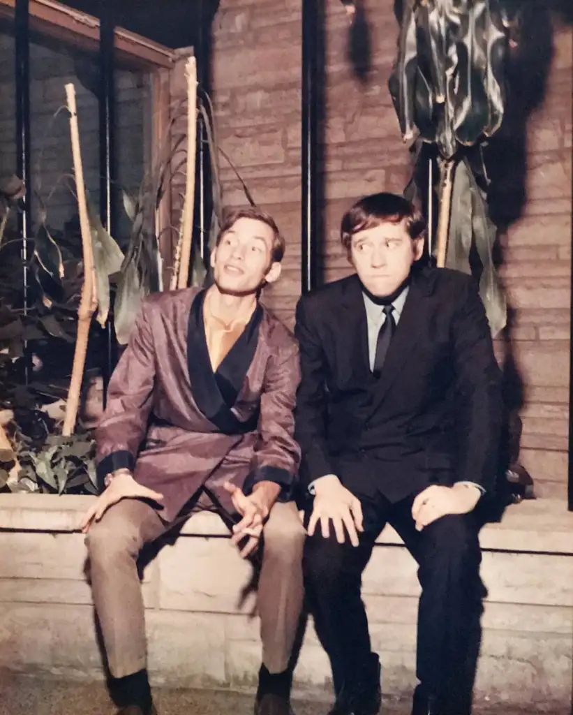 Two men are sitting on a low stone wall indoors. The man on the left wears a satin robe and light pants, gesturing animatedly, while the man on the right in a dark suit listens, looking amused. Tall plants and wood-paneled walls form the background.