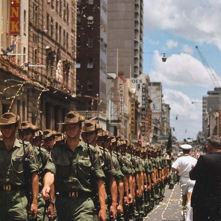 A parade of soldiers in green uniforms and hats march down a city street. Confetti streams from above. Tall buildings line the street, and spectators watch from the sidewalk. The sky is partly cloudy.