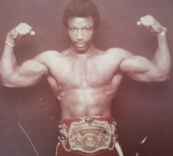 A muscular man confidently poses with his arms flexed, displaying a championship belt around his waist. The individual has a serious expression and stands against a plain background.