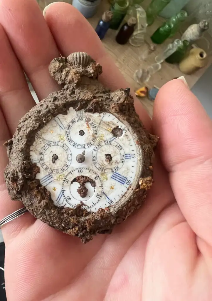 A hand holds an old, rusty pocket watch with a cracked face. The watch is covered in dirt and corrosion, and the time is unclear. In the background, there are various glass bottles and jars on a wooden shelf.