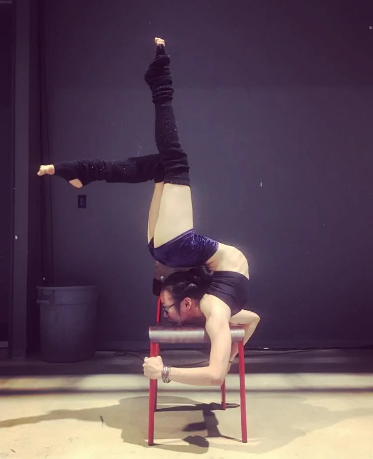 A person performs a balancing yoga pose on a chair with their legs elevated and one leg bent. They wear a black top, dark leg warmers, and purple shorts. The room has a dark wall and there's a small trash bin in the background.