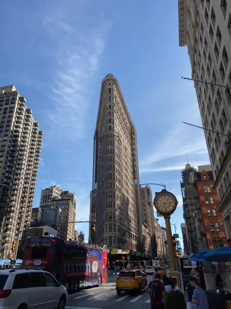 Street view of a bustling city with the Flatiron Building prominently in the center. Surrounding buildings rise alongside, with a mix of traffic including a tour bus and taxis. A vintage street clock stands on the right under a clear blue sky.