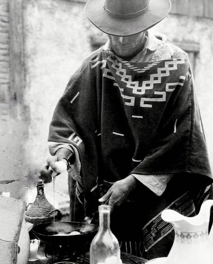 Black and white image of a person wearing a wide-brimmed hat and patterned poncho, cooking eggs in a pan outdoors. Various bottles and a pitcher are on the table. The setting appears rustic.