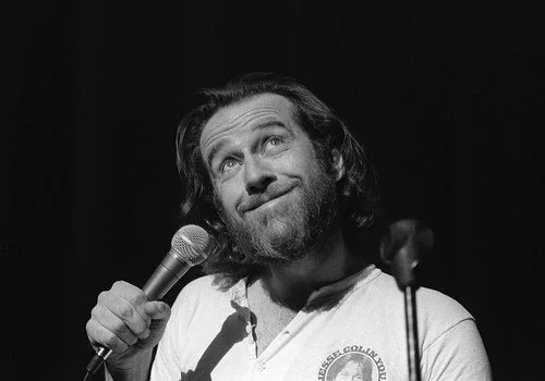 A bearded man with long hair stands on stage, holding a microphone. He looks upward with a thoughtful expression. He's wearing a light-colored shirt featuring a graphic. The image is in black and white.