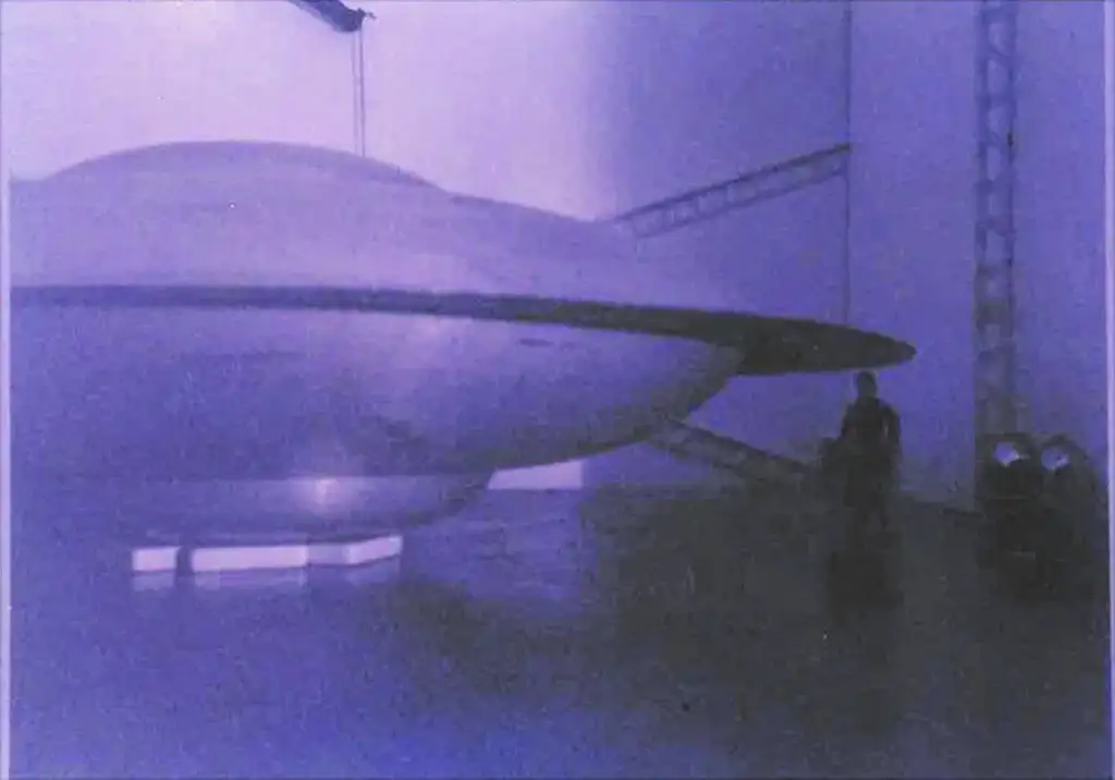 A large, disc-shaped UFO is displayed in an indoor setting, bathed in blue light. A person stands nearby for scale, highlighting the massive size of the object. The space appears to be a hangar or exhibit area.