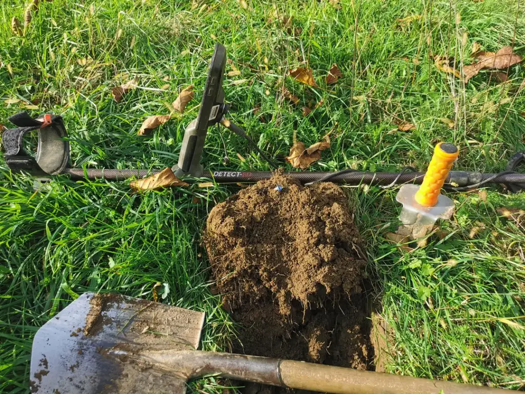 Metal detecting scene: A metal detector rests on grass beside a mound of unearthed soil. A small hand shovel is nearby, partially buried. A pinpointing tool with an orange handle is also present. Scattered leaves add autumnal touches.