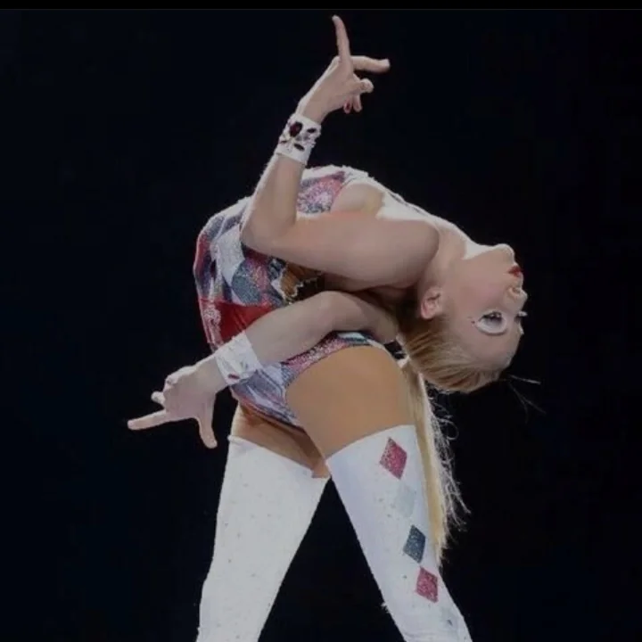 A performer in a colorful costume strikes an acrobatic pose against a dark background. They are bending backwards with one arm extended behind and the other across the chest, showcasing flexibility and balance.