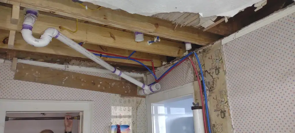 Visible ceiling damage with exposed wooden beams and plumbing pipes. Wallpapered walls show floral patterns, and part of the ceiling is peeling. A partially visible door and window are in the background.