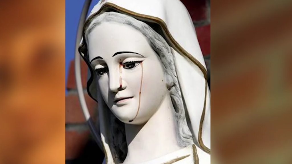 A statue of the Virgin Mary appears to be shedding tears of blood. The white statue has a serene expression, with the red streaks vividly visible against her face. Background is blurred, emphasizing the striking image.