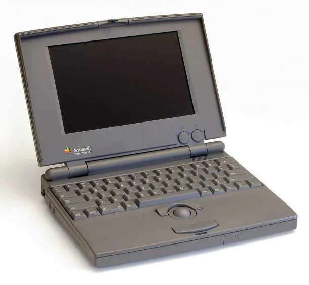 A vintage gray laptop with an open screen, displaying a blank black display. It has a QWERTY keyboard and a built-in trackball. The Apple logo is visible near the display hinge.