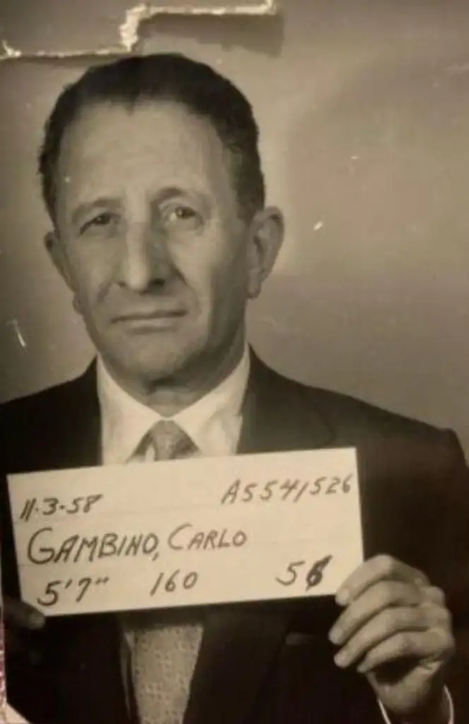 A black and white mugshot photo of a man in a suit holding a board with handwritten information: "11-3-58,  AS541526, Gambino, Carlo, 5'7", 160, 56." The man looks directly at the camera with a neutral expression.