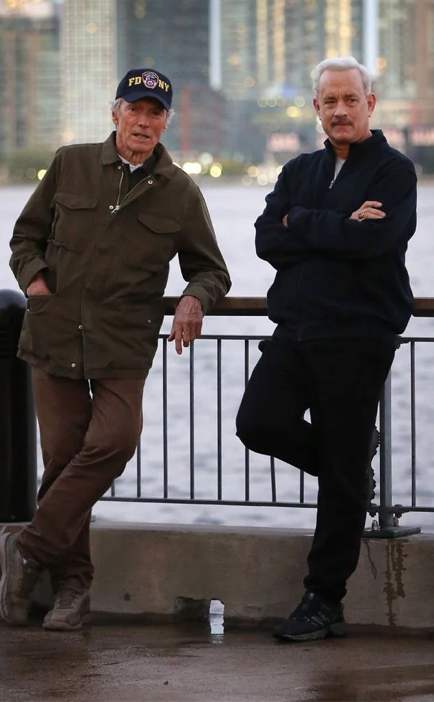 Two men stand by a waterfront railing with a city skyline in the background. The older man on the left wears a cap and green jacket, while the younger man on the right wears a black jacket and has his arms crossed.