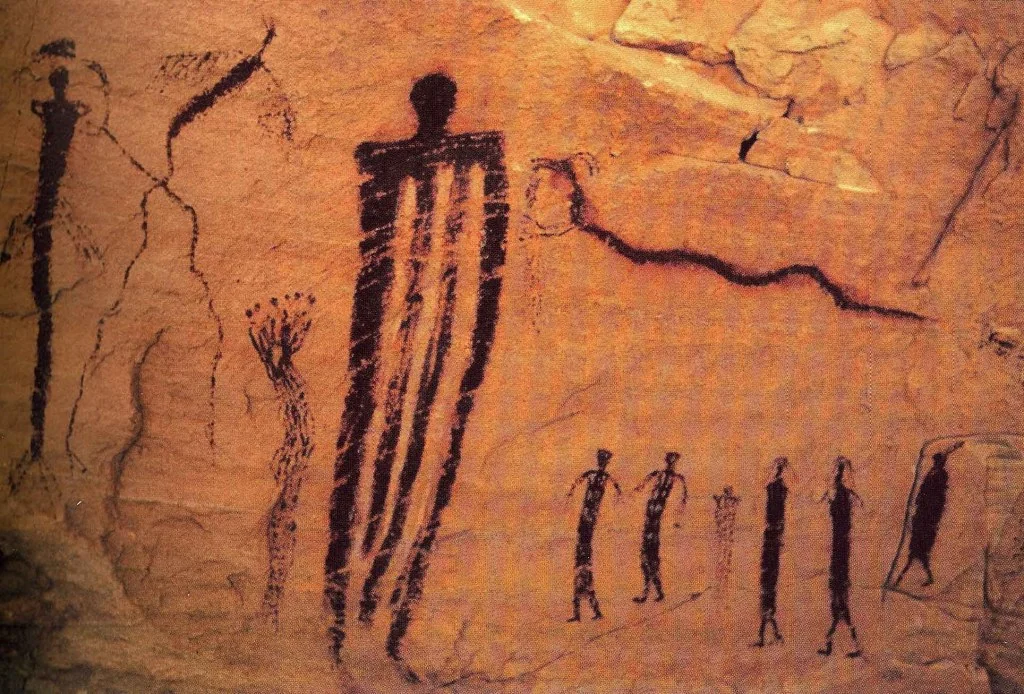 Ancient rock art on a reddish-brown stone surface depicting silhouettes of human-like figures with intricate patterns. The main central figure is taller and more prominent than the others, surrounded by smaller figures and abstract line designs.