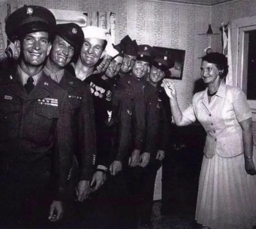 A black and white photo showing five men in military uniforms, posing in a line. They are smiling and standing close together. A woman in a dress gestures toward them with a smile in a room with patterned wallpaper.