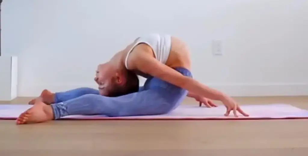 A person is performing a yoga pose on a pink mat. They are bending backward with their head touching the ground and arms extended, while seated with legs open. They are wearing a white top and blue leggings in a bright room.