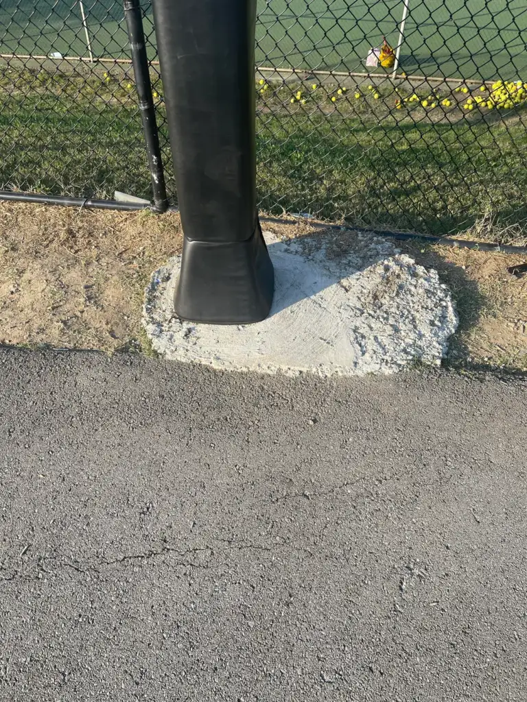 A field with a chain-link fence and a padded black post. The ground is paved with asphalt and a patch of concrete at the base of the post. Yellow flowers and green grass can be seen in the background.